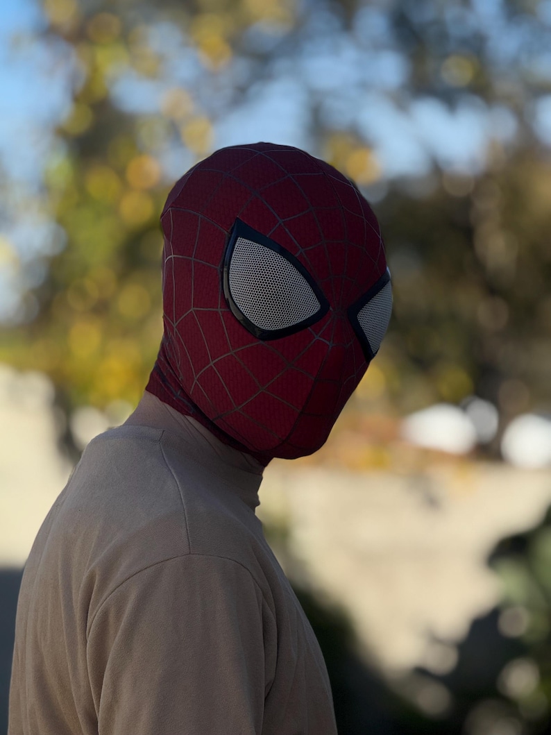 Puede incluir: Una m&aacute;scara de Spider-Man roja y negra con cubiertas oculares de malla blanca. La m&aacute;scara tiene un patr&oacute;n de telara&ntilde;a y se usa sobre la cabeza. La persona lleva una camiseta de manga larga de color beige.