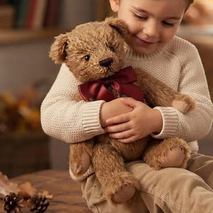 Könnte beinhalten: Ein brauner Plüsch-Teddybär mit einer dunkelroten Satinschleife um den Hals. Der Bär hat strukturiertes Fell, gestickte Gesichtsdetails und Tatzen aus hellbraunem Stoff. Er wird in Armen gehalten und zeigt seine weiche, ausgestopfte Konstruktion.