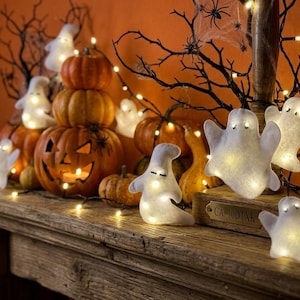 May include: A collection of white felt ghost ornaments illuminated by internal warm LED lights, arranged alongside orange pumpkins and a carved jack-o'-lantern on a wooden mantelpiece. The display features bare branches and string lights.