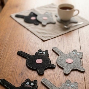 May include: Felt cat-shaped coasters in black and gray, with pink circles and white stitching. The coasters are on a wooden table, with a cup of tea and a linen napkin in the background. The text "HANDARBEIT & CHARAKTER - 100% FILZ" is at the bottom.