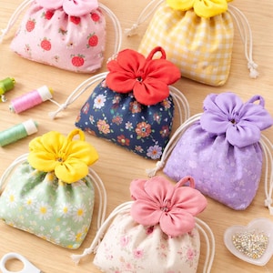 Könnte beinhalten: Eine Sammlung von Beuteln mit Kordelzug in verschiedenen floralen und gemusterten Stoffen, die jeweils mit einer großen Stoffblume verziert sind. Farben sind Rosa, Gelb, Rot, Blau und Lila. Weiße Kordelzüge und eine Schere sind ebenfalls zu sehen.