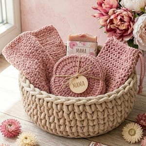 May include: A woven basket filled with pink crocheted items, including washcloths and coasters, and a bar of soap. A wooden tag with "MAMA" is attached. A card with text and flowers are also present.