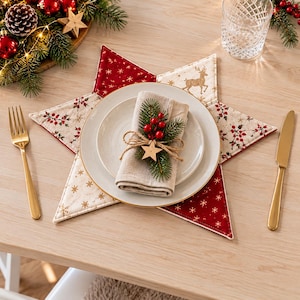 May include: A star-shaped fabric placemat featuring red and cream panels with gold reindeer and snowflake patterns. A beige linen napkin is folded on a white plate, adorned with a sprig of pine, red berries, and a small wooden star.