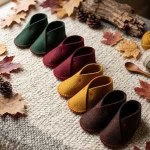 Könnte beinhalten: Vier Paar Babyschuhe aus gefilzter Wolle, angeordnet in einer diagonalen Linie, in den Farben Dunkelgrün, tiefes Burgunderrot, Senfgelb und Dunkelbraun. Jedes Paar hat ein einfaches, überlappendes Design mit kleinen roten Kreuzstichen.