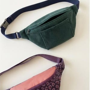 May include: Two bum bags. One is dark green corduroy with a navy blue adjustable strap and a front zipper pocket. The second bum bag features a purple leopard print pattern with a solid pink top panel, a purple zipper, and a purple strap.