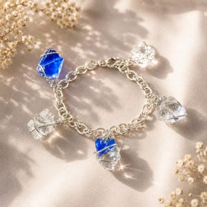 Könnte beinhalten: Ein silbernes Charm-Armband mit klaren und blauen Kristall-Anhängern. Das Armband hat ein Kettenglied-Design mit mehreren Kristall-Anhängern. Die Kristalle sind in Silberdraht gewickelt. Der Hintergrund ist eine neutrale Farbe mit weicher Beleuchtung.