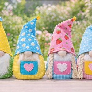 Könnte beinhalten: Vier dekorative Zwerge mit Stoffhüten und -körpern. Die Hüte sind gelb mit weißen Punkten, blau mit weißen Gänseblümchen, rosa mit Erdbeeren und blau mit weißen Gänseblümchen. Jeder Zwerg hat einen weißen Bart und eine herzförmige Schürze.