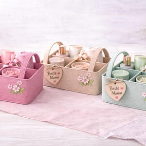 Könnte beinhalten: Drei Filz-Caddies in Rosa, Beige und Mintgrün, jeweils mit Griff und unterteilten Fächern. Sie enthalten Schönheitsprodukte und sind mit floralen Akzenten und einem Holzherzanhänger mit der Aufschrift "Beste Mama" verziert.
