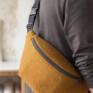 May include: A mustard yellow felted crossbody bag featuring a curved top zip closure with a dark grey metal pull. The bag is attached to an adjustable black nylon strap with a matching dark grey plastic side-release buckle and slider hardware.