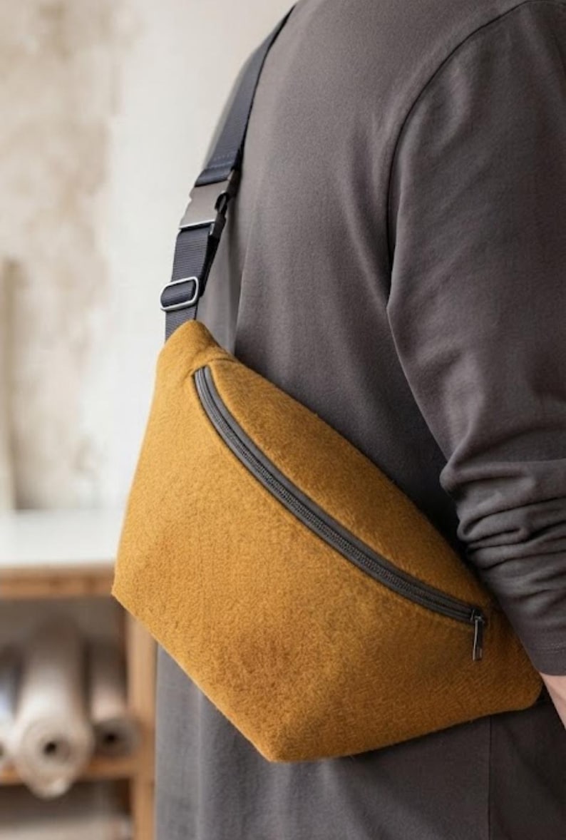 May include: A mustard yellow felted crossbody bag featuring a curved top zip closure with a dark grey metal pull. The bag is attached to an adjustable black nylon strap with a matching dark grey plastic side-release buckle and slider hardware.
