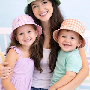 Könnte beinhalten: Eine Frau und zwei Kinder tragen passende Fischerhüte. Die Hüte sind in Pastellfarben und Mustern gehalten, darunter ein grünes Blumenmuster, rosa Vichy-Karo und orangefarbenes Vichy-Karo. Die Frau trägt ein weißes Oberteil und Jeans.