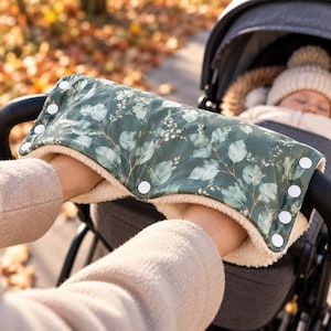 May include: A stroller hand muff featuring a dark green exterior with a white and green botanical leaf pattern. The interior is lined with cream-colored faux fleece. The muff attaches to the stroller handle with white plastic snap buttons.