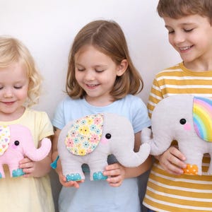 Op de afbeelding: Drie kinderen houden pluche olifantenspeelgoed vast. De olifanten zijn roze, grijs en grijs met regenboogaccenten. Elke olifant heeft een andere oor met patroon en kleurrijke voeten. De kinderen glimlachen.