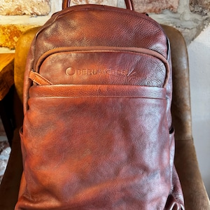 May include: A brown leather backpack with a top handle and multiple pockets. The backpack has a front pocket with a zipper and the brand name "BERLAZIDELE" embossed on it. The leather has a distressed look, and the backpack is sitting on a brown chair.