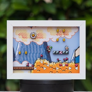 May include: A rectangular white shadow box frame containing a multi-layered paper diorama of a video game scene. The artwork features a blue sky, a large mountain, orange foliage, floating gold coins, a flower, and pixelated character figures.