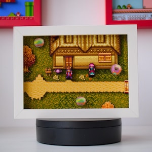 Op de afbeelding: Een ingelijst pixel art diorama met een scène uit een videogame. Het kunstwerk toont een huis, personages en een pad op een groen landschap. De lijst is wit en het diorama wordt gepresenteerd op een zwarte draaiende standaard. Twee andere ingelijste kunstwerken zijn zichtbaar op de achtergrond.