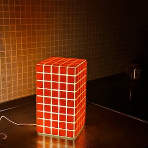 Può includere: Una lampada rossa a forma di cubo con un motivo a griglia di piccoli quadrati. La lampada è illuminata dall'interno, proiettando un bagliore caldo. La lampada è su una superficie scura, con un cavo di alimentazione bianco.