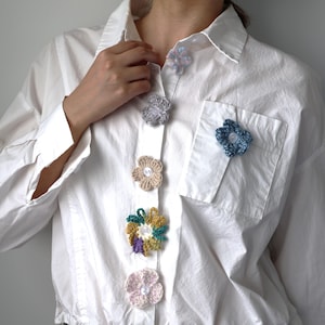 May include: A white shirt featuring a series of crocheted flower buttons in diverse colours. The handmade floral decorations are attached to the shirt, including a pocket pin, offering a unique, handcrafted aesthetic.