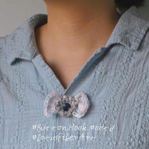 May include: A crocheted bow-shaped button with a dark blue centre button, attached to a light blue collared shirt. The bow is grey, pink, and white. The text "#ButtonHook #ootd #LotusFiberArt" is at the bottom.