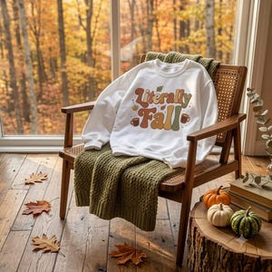 Op de afbeelding: Een wit sweatshirt met lange mouwen en de tekst "Literally Fall" in een retro, meerkleurig lettertype. De afbeelding bevat kleine illustraties van herfstbladeren en koffiemokken in tinten oranje, bruin, geel en groen.