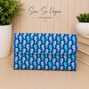May include: A rectangular fabric wallet featuring a repeating pattern of seahorses in shades of blue, teal, and yellow against a dark blue background. The wallet has a fold-over flap closure and a structured design with visible stitching along the edges.