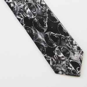 May include: A black and white patterned necktie featuring a repeating design of coffee bean shapes. The tie has a diagonal orientation against a white background. The pattern is a mix of grayscale tones, creating a textured appearance.