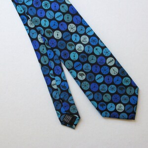 May include: A blue silk tie with a repeating pattern of blue and teal circles on a black background. Each circle contains a unique, abstract design. The tie is angled across a white surface, showcasing its design and texture.