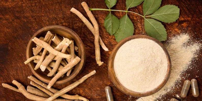 K&ouml;nnte beinhalten: Draufsicht auf Ashwagandha-Produkte. Zwei Holzschalen enthalten Ashwagandha-Wurzelst&uuml;cke und -Pulver. Kapseln, Bl&auml;tter und Wurzelst&uuml;cke sind auf einer Holzoberfl&auml;che verstreut.