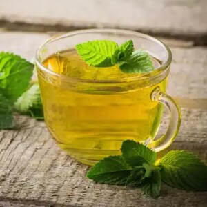 May include: A clear glass teacup filled with golden-yellow tea, garnished with fresh green mint leaves. The cup sits on a weathered wooden surface, with additional mint leaves scattered around. The tea appears warm and inviting.