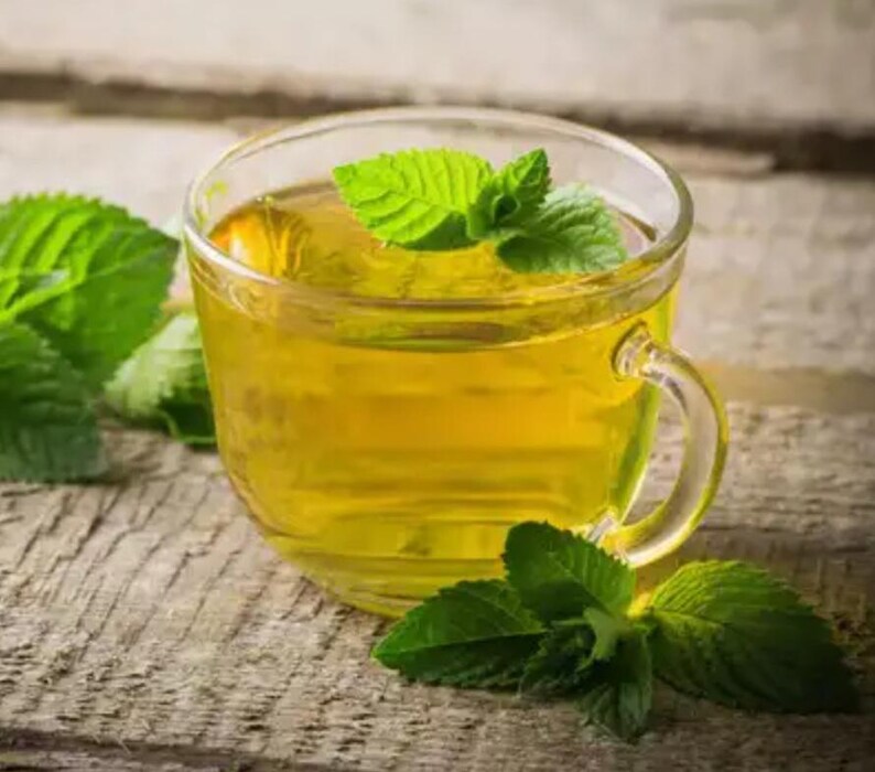 May include: A clear glass teacup filled with golden-yellow tea, garnished with fresh green mint leaves. The cup sits on a weathered wooden surface, with additional mint leaves scattered around. The tea appears warm and inviting.