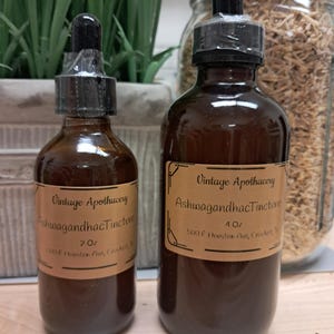 K&ouml;nnte beinhalten: Zwei braune Glasflaschen mit schwarzen Tropfern und Etiketten mit der Aufschrift "Vintage Apothecary Ashwagandha Tincture". Die Flaschen sind 60 ml und 120 ml. Der Hintergrund umfasst Gr&uuml;n und ein Glas mit getrockneten Kr&auml;utern.