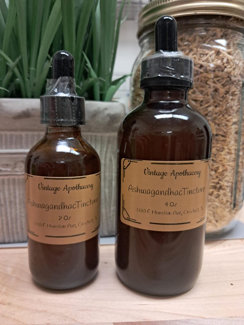 K&ouml;nnte beinhalten: Zwei braune Glasflaschen mit schwarzen Tropfern und Etiketten mit der Aufschrift "Vintage Apothecary Ashwagandha Tincture". Die Flaschen sind 60 ml und 120 ml. Der Hintergrund umfasst Gr&uuml;n und ein Glas mit getrockneten Kr&auml;utern.