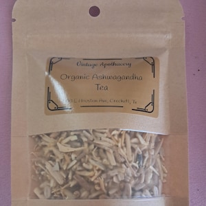 May include: A tan pouch with a clear window containing dried herbs. The label reads "Vintage Apothecary Organic Ashwagandha Tea." The pouch is sealed and has a small hole at the top. The background is a light purple.