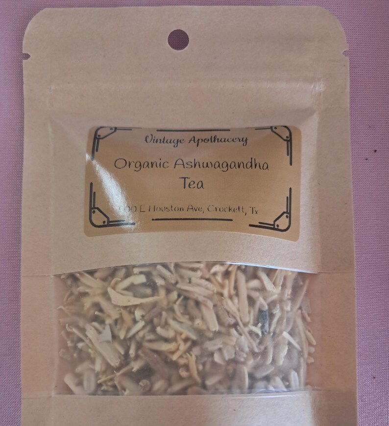 May include: A tan pouch with a clear window containing dried herbs. The label reads "Vintage Apothecary Organic Ashwagandha Tea." The pouch is sealed and has a small hole at the top. The background is a light purple.