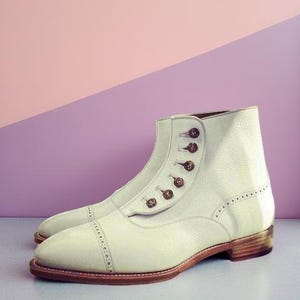 Puede incluir: Un par de botines de cuero blanco roto con puntera y cierre de botones. Las botas tienen un tacón apilado y suela marrón. El fondo es una división de rosa y morado.