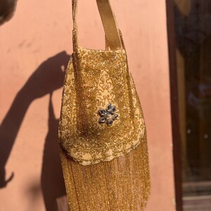 May include: A vintage gold beaded handbag with a long handle. The bag is covered in small, shimmering beads and features a decorative floral embellishment. The bottom of the bag has a fringe of gold beads.