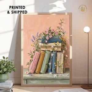 May include: A framed art print featuring a stack of books on a table, adorned with climbing pink and yellow flowers and lavender sprigs. The background is a solid peach color, and the books are rendered in shades of pink, yellow, green, and blue.