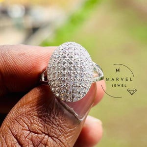 Puede incluir: Un anillo de plata con una parte superior de forma ovalada cubierta de pequeñas piedras brillantes. El anillo se sostiene contra un fondo borroso de follaje verde y un edificio. El anillo es una joya.