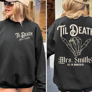 May include: Black sweatshirt with the text "Til Death Mrs. Smith" on the front and back. The back features a skeleton hand with a ring. The date III.IV.MMXXVI is printed below the name. The sweatshirt is black.