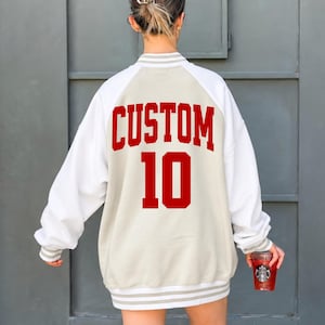 May include: A beige and white varsity-style jacket featuring long white sleeves with ribbed cuffs and a beige body. The back displays the word CUSTOM in bold red capital letters above the number 10 in a matching red font with a white outline.