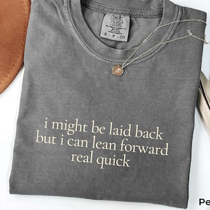 以下が含まれることがあります： 「i might be laid back but i can lean forward real quick」という白文字がプリントされたグレーのTシャツ。金のネックレスと茶色の帽子も写っています。シャツには「Pepper」と書かれています。