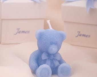 May include: A light blue teddy bear candle with a bow, sitting in front of white gift boxes tied with light blue ribbons. The boxes have the name "James" printed on them. The candle is a charming decorative item.