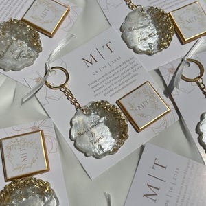 Könnte beinhalten: Schlüsselanhänger mit goldener Kette und Ring, mit einem weißen und goldenen Harzdesign. Jeder Schlüsselanhänger wird mit einer Karte mit den Initialen "M|T" und dem Datum "06/16/2025" geliefert. Die Namen "Mohammad & Tayma" sind auf dem Schlüsselanhänger geschrieben.