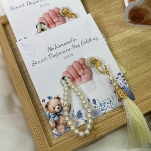 May include: Two cards with the text "Muhammed'in Sünnet Düğününe Hoş Geldiniz" and the date 11.10.25. Each card features a baby's hand holding a pearl rosary and a gold-colored charm. A small teddy bear illustration is also present.