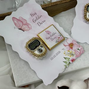 May include: A decorative favor featuring a digital tally counter with a pearl and gold-tone border, a small chocolate bar, and a card with floral and butterfly designs. The card has the text "Hoş Geldin Duru" and the date 03.03.2026.