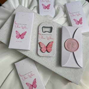 Pode incluir: Uma coleção de lembrancinhas de chá de bebê, incluindo um abridor de garrafas e pequenas caixas. Os itens são brancos com desenhos de borboletas rosa e texto. O abridor de garrafas tem um design de borboleta rosa. As caixas são seladas com um selo de cera rosa.