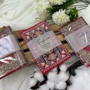 May include: Red and gold patterned fabric pouches with a gold cord loop. Each pouch is wrapped with a translucent paper band with text. The pouches are arranged on a wooden tray with white flowers in the background.