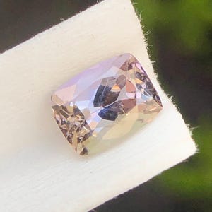 May include: A faceted, rectangular gemstone with a soft, iridescent color palette of lavender, peach, and pale yellow. The gem is set on a white, textured surface, with a dark background.