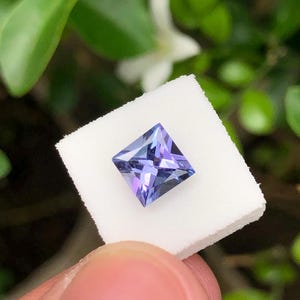 以下が含まれることがあります： 鮮やかな紫と青のカラーパレットが特徴のスクエアカットの宝石。光を反射する多面的な表面を持ち、小さな正方形の白いフォームディスプレイパッドに固定されており、その幾何学的な形状がはっきりと示されています。