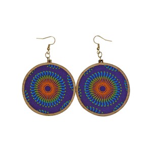 May include: A pair of round earrings with a purple fabric design featuring an orange and blue circular pattern. The fabric is set in a wooden frame with gold-colored hooks. The earrings are a unique accessory.